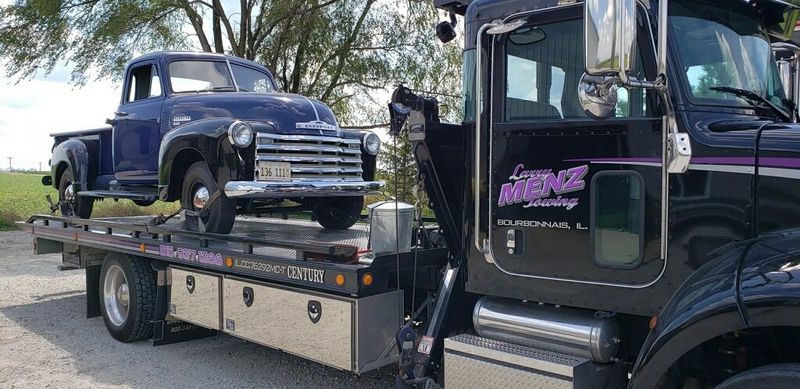 Black Larry Menz wrecker with purple Bourbonnais door lettering hauling a vintage Chevrolet pickup