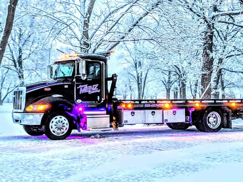 Black Larry Menz Peterbilt flatbed on a snow-covered Illinois road, amber lights on